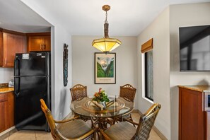 A compact kitchen with a black refrigerator, wooden cabinets, a dining table with wicker chairs, and a hanging light fixture. - Kihei Beach, #505 1 Bedroom Condo (Kihei)