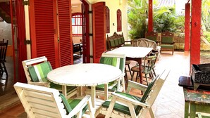 Quarto com mesa redonda e cadeiras, persianas vermelhas e vista para o verde lá fora.