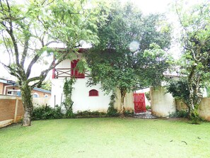 A white building with red accents, surrounded by a grassy yard and trees. - Casa com muito lazer a 100m da Praia de Guaecá/SP (São Sebastião)