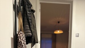 A hallway with a coat rack holding a leopard print bag and a black jacket. A shoe rack with sneakers is visible. The hallway leads to a living area with a couch and a TV. - Family-friendly and cozy guesthouse on the edge of the forest. (Kasterlee)