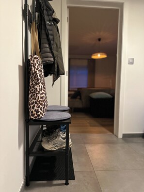 A hallway with a coat rack holding a leopard print bag and a black jacket. A shoe rack with sneakers is visible. The hallway leads to a living area with a couch and a TV. - Family-friendly and cozy guesthouse on the edge of the forest. (Kasterlee)