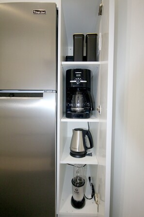 Eine kompakte Küche mit integriertem Kühlschrank, einer Kaffeemaschine, einem Wasserkocher und einem Mixer auf einem weißen Regal.