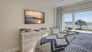 Ein Schlafzimmer mit großem Fenster und Blick auf den Strand, einem Bett mit gemustertem Überwurf, einer Kommode mit Dekoration und einem gerahmten Bild an der Wand.