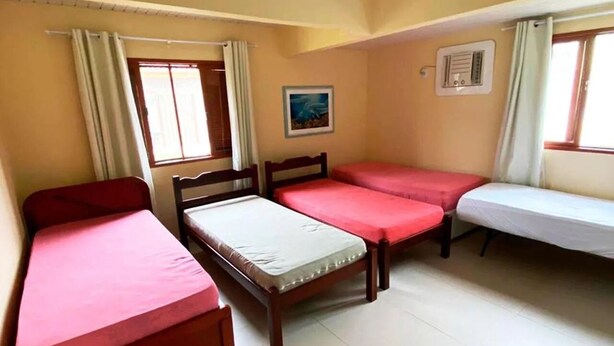 A dormitory room with three single beds, a window with curtains, a radiator, and a painting on the wall. - Imperdível: Incrível casa a 65m da praia do Meaípe (Guarapari)