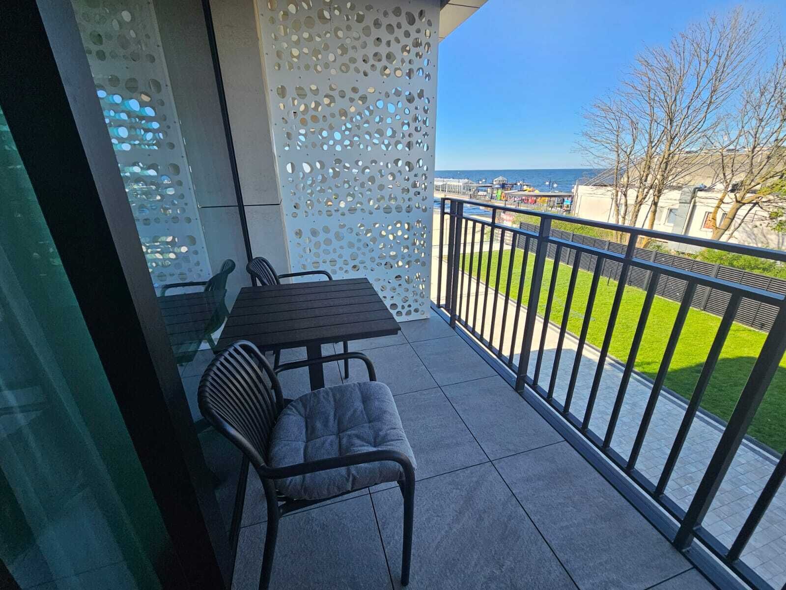 Un balcón con mesa y sillas, un muro decorativo perforado y vistas al mar y vegetación.