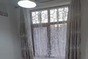 A room with a large window covered by floral curtains, a ceiling light fixture, and a wall-mounted air conditioning unit. - Family- Near Lcy Airport- Sleeps 6- Parking- Wi-fi (Dagenham)