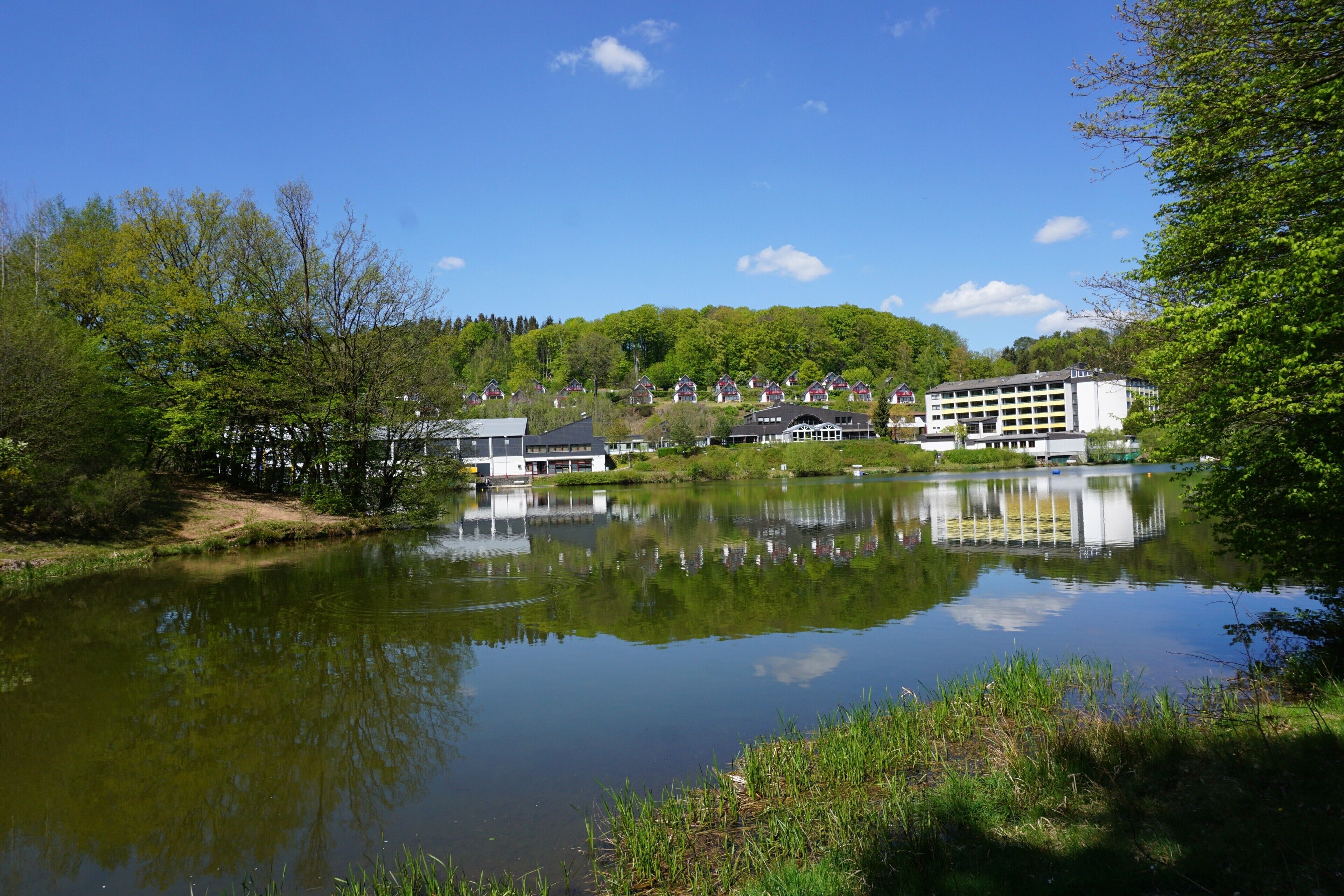 A lakeside hotel with a reflection in the water, surrounded by greenery and trees.