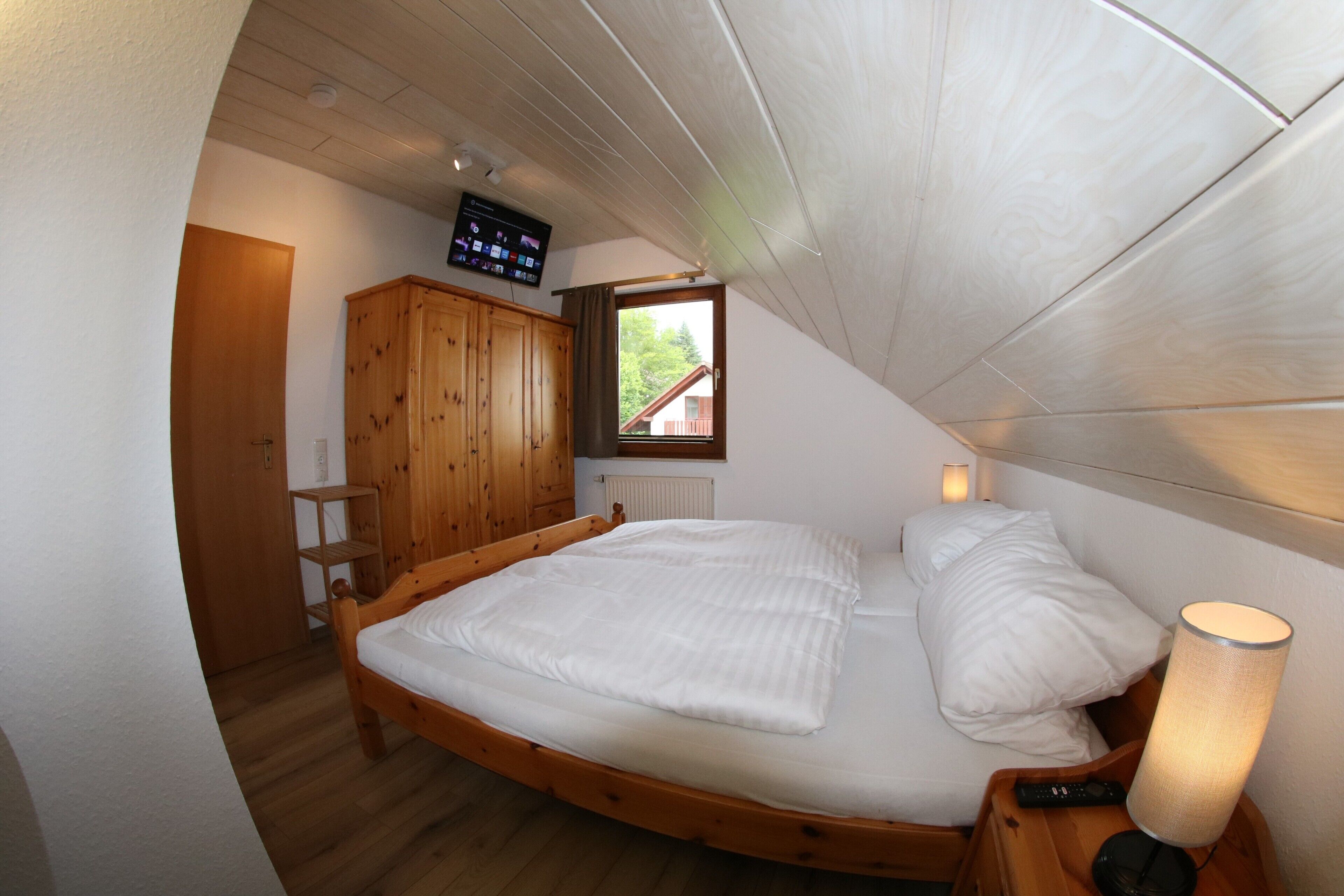 Ein Schlafzimmer mit einem hölzernen Bett, einem Nachttisch, einer Lampe, einem Holzschrank, einem an der Wand befestigten Fernseher und einem Fenster mit Blick auf ein Haus.