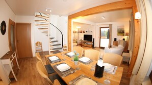 Uma sala de jantar com mesa de madeira posta para quatro pessoas, uma escada em espiral e uma sala de estar com sofá e televisão.