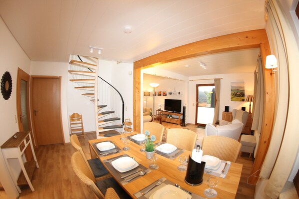 Un coin repas avec une table en bois dressée pour quatre personnes, un escalier en colimaçon et un salon avec un canapé et un téléviseur.