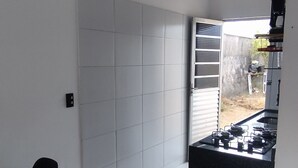 Eine kompakte Küche mit Gaskochfeld und einer kleinen Arbeitsfläche. Ein Essbereich mit Holztisch und gelben Stühlen ist ebenfalls vorhanden. Der Raum hat weiße Wände und einen gefliesten Boden.