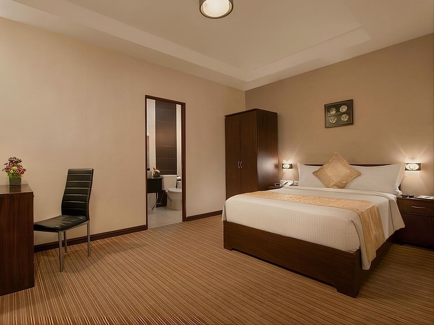 Una habitació d'hotel amb un llit gran, un armari de fusta, un escriptori amb una cadira i un bany visible a través d'una porta oberta.