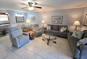 Un salon avec un canapé gris, un fauteuil bleu, une table basse en verre et un ventilateur de plafond.