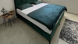 Ein Schlafzimmer mit einem grünen Bett, weißer Bettwäsche, einem kleinen Holfnachtkästchen und einem Fenster mit Blick auf Grünflächen.