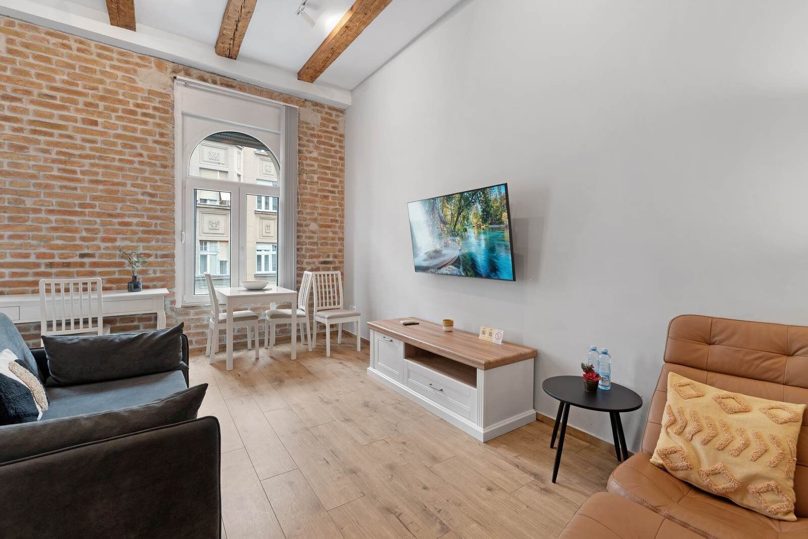 A modern living room with a brick wall, a dining area with white furniture, a TV mounted on the wall, and a sofa with a cushion.