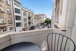 A balcony with a view of a street lined with buildings, a chair, and a table.