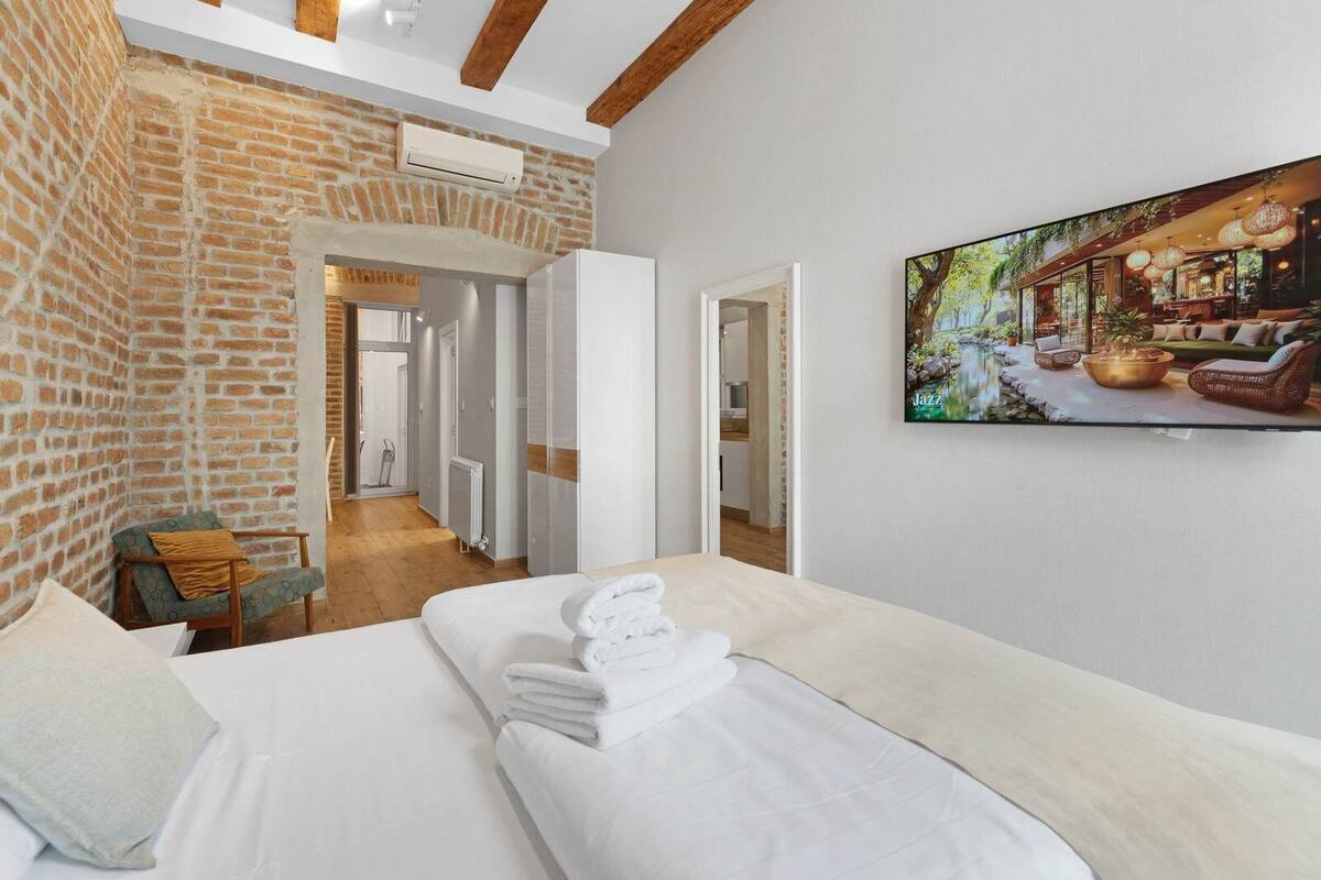 Una habitación de hotel con pared de ladrillo, una cama con ropa blanca, una silla y un cuadro de un jardín.