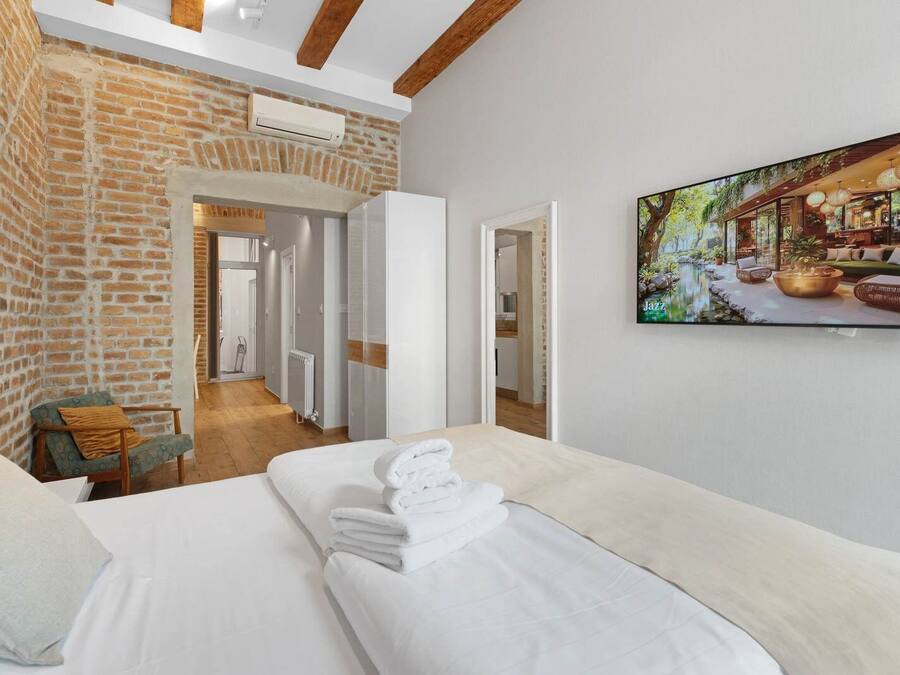A hotel room with a brick wall, a bed with white linens, a chair, and a painting of a garden scene.