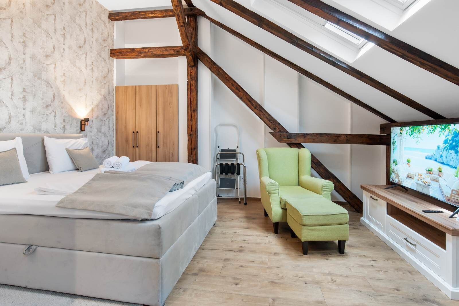 Een slaapkamer met een bed, een groene fauteuil, een televisie en een houten garderobe.