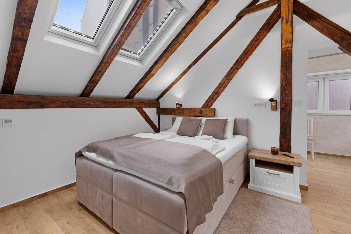 Un dormitorio con techo inclinado, vigas de madera, cama, mesita de noche y ventana.
