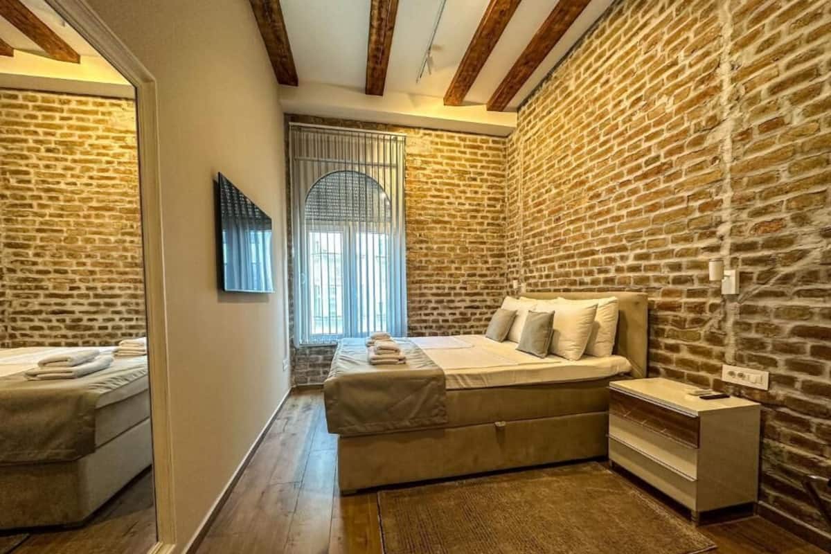 Un dormitorio con una cama grande, una cama más pequeña, una mesita de noche, un espejo y una ventana con persianas.