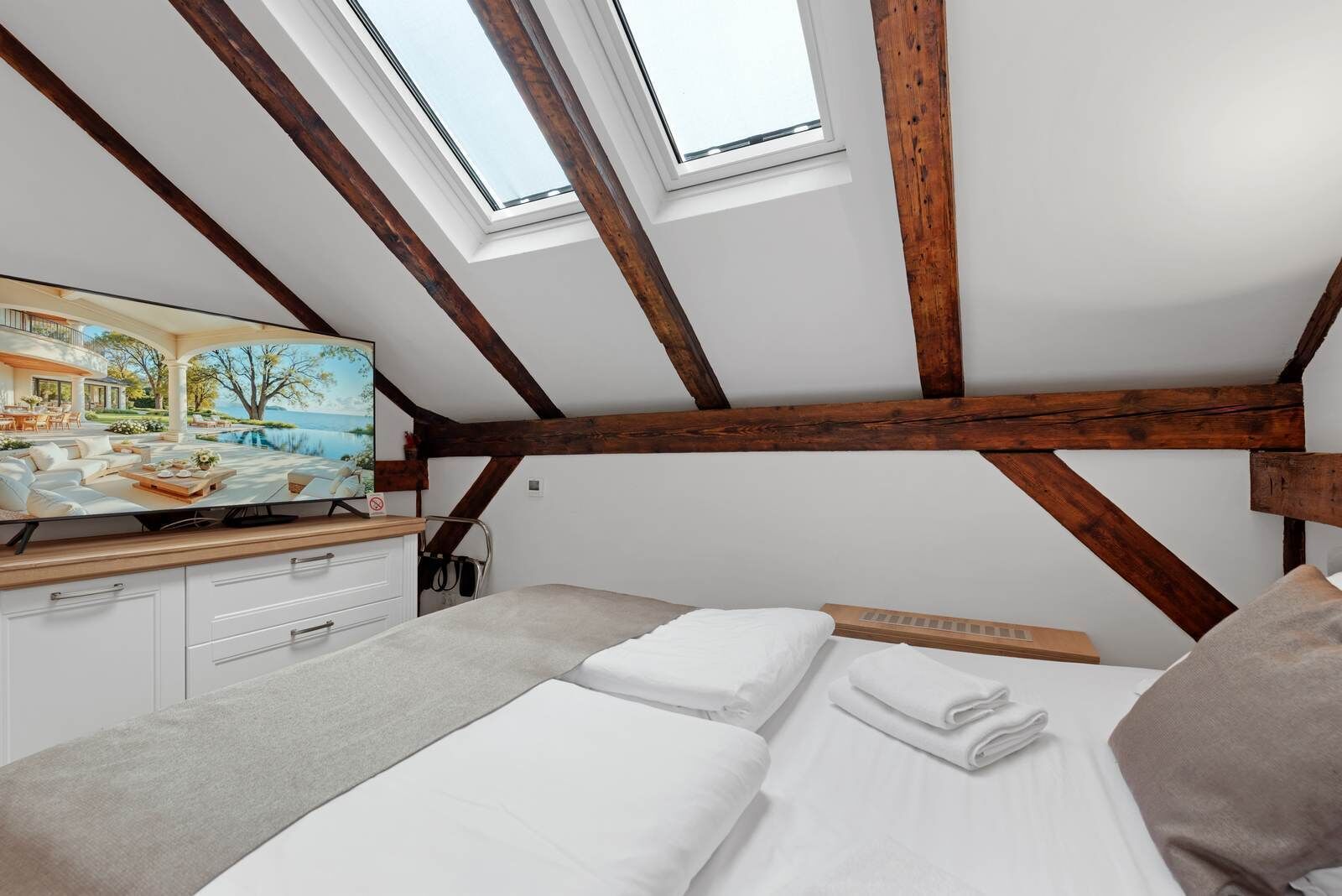 A bedroom with a sloped ceiling, wooden beams, a bed, a TV, and a view of a house and water outside.