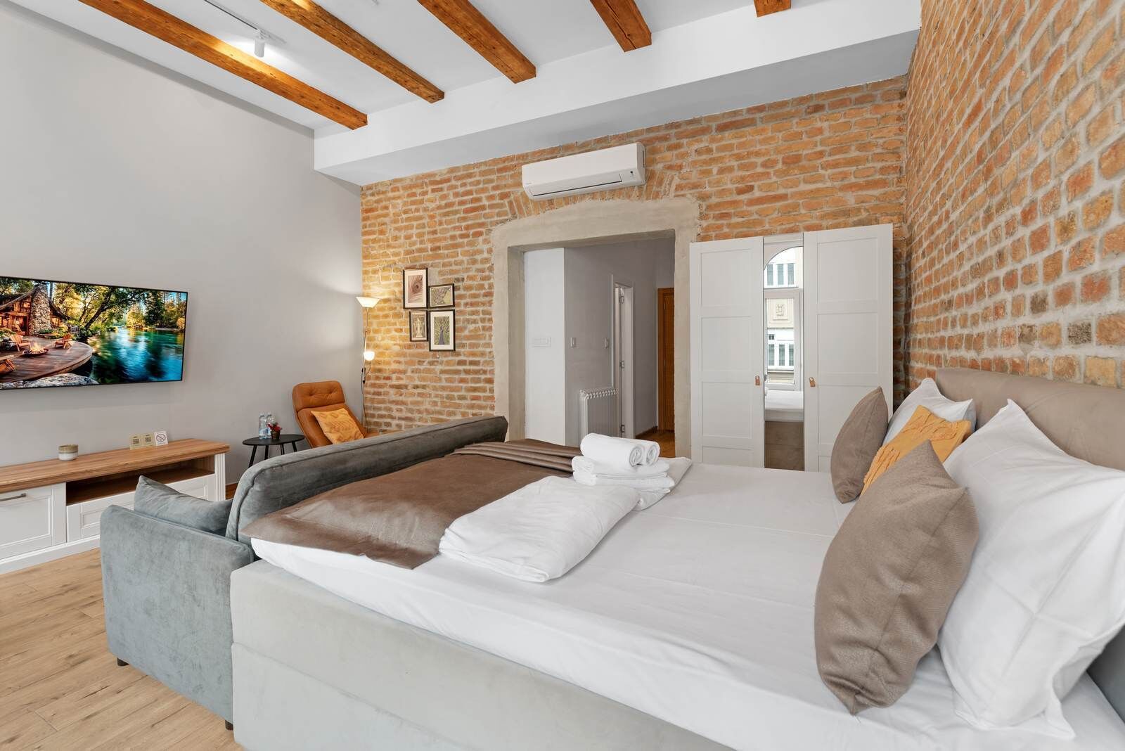 A modern hotel room with a brick wall, a large bed with white linens, a TV mounted on the wall, and a cozy seating area with a chair and a small table.