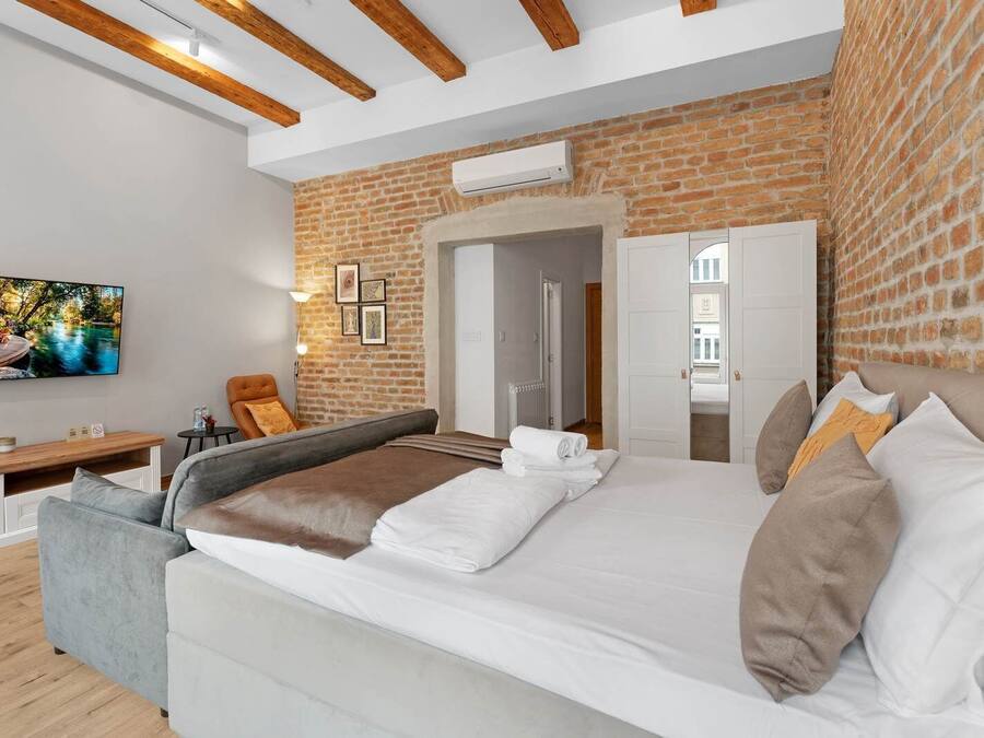A modern hotel room with a brick wall, a large bed with white linens, a TV mounted on the wall, and a cozy seating area with a chair and a small table.