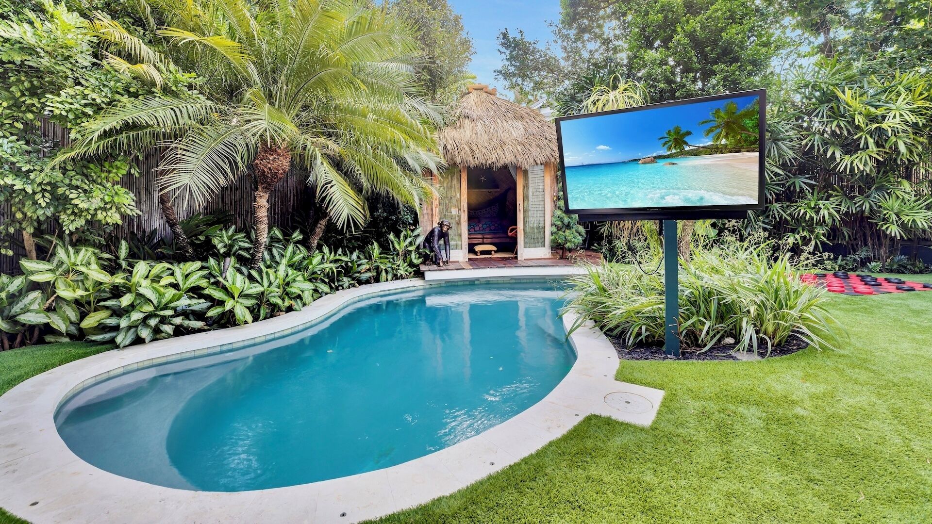 A swimming pool surrounded by lush greenery, a thatched-roof structure, and a large screen displaying a tropical beach scene.