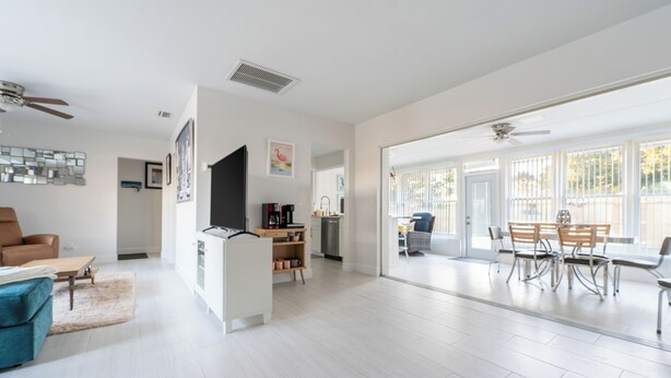 A spacious living area with a sofa, coffee table, and a dining table with chairs. There is a kitchen area with cabinets and a refrigerator. The room has large windows and a ceiling fan. - Charming 3-bedroom house with AC in Sunny Venice... High End Pool/Fenced in yard (Venice)