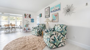 A living room with two matching armchairs, a fluffy rug, a dining area with a table and chairs, and wall art including a letter 'S' and travel-themed pictures. - Charming 3-bedroom house with AC in Sunny Venice... High End Pool/Fenced in yard (Venice)