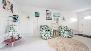 A living room with a red coffee table, two floral armchairs, a shaggy rug, and wall art including a clock and framed pictures. - Charming 3-bedroom house with AC in Sunny Venice... High End Pool/Fenced in yard (Venice)