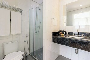 A bathroom with a glass-enclosed shower, a white toilet, a sink with a black countertop, and a mirror above the sink. - St. Charbel by Charlie (São Paulo)