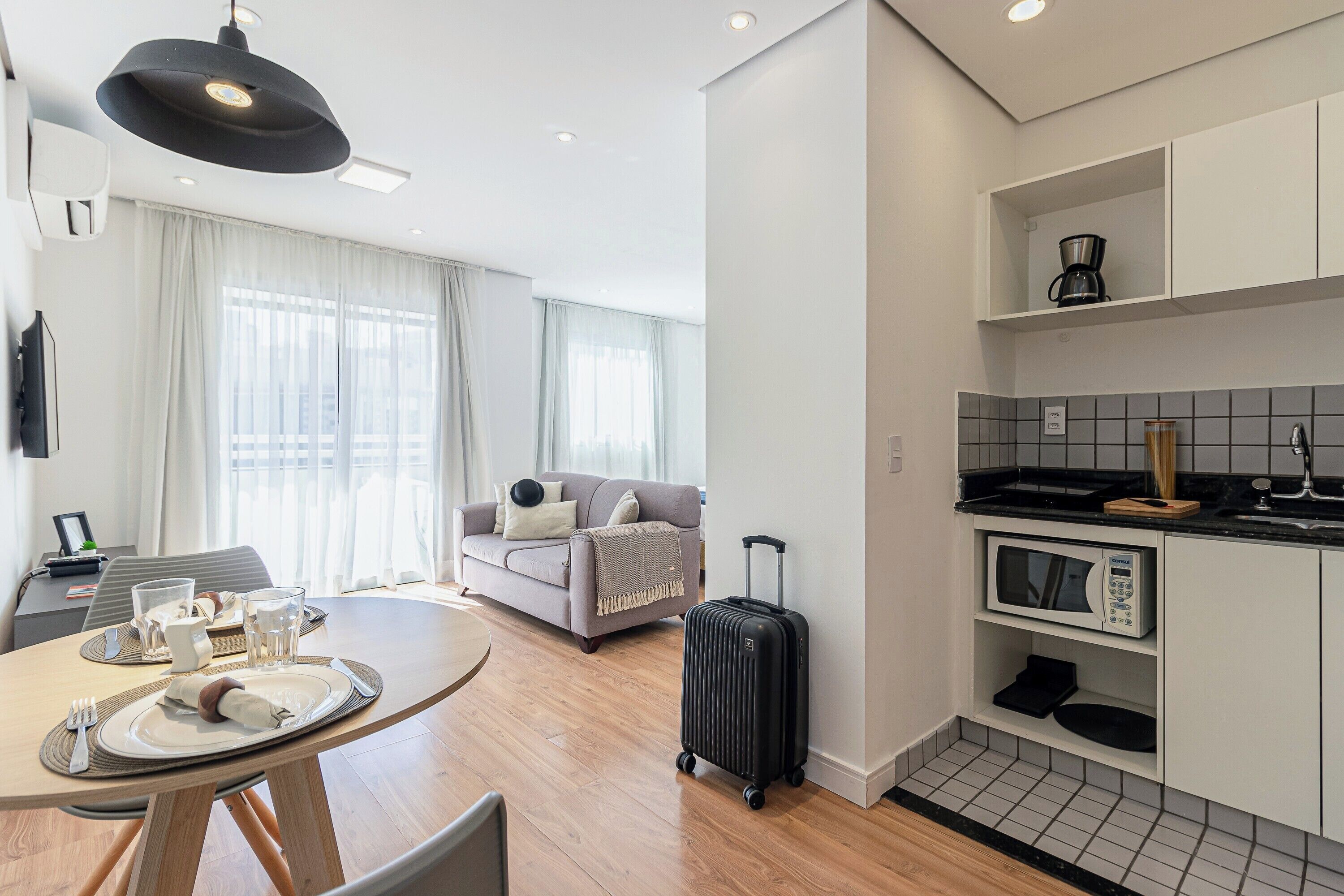A modern hotel room with a dining area, a sofa, a kitchenette, and a suitcase.