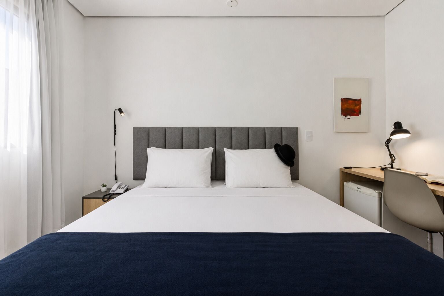 A hotel room with a bed, bedside table, lamp, chair, and a small desk with a lamp.
