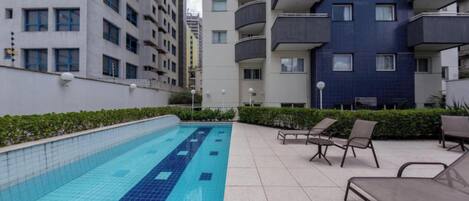 A swimming pool with tiled flooring, surrounded by lounge chairs and a low hedge.