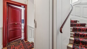 Un escalier recouvert de moquettes à motifs rouges et une porte rouge sur laquelle est posé un paillasson avec l’inscription « BIENVENUE ».