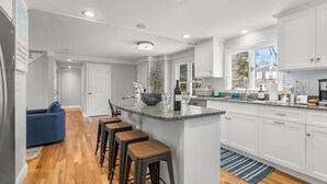 A modern kitchen with white cabinets, a granite countertop, and a central island with stools. - Falmouth Family Home: Beach, Fire Pit & Games! (Falmouth)