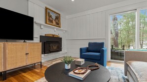 A living room with a fireplace, a television, and a large window with a view of trees. - Falmouth Family Home: Beach, Fire Pit & Games! (Falmouth)