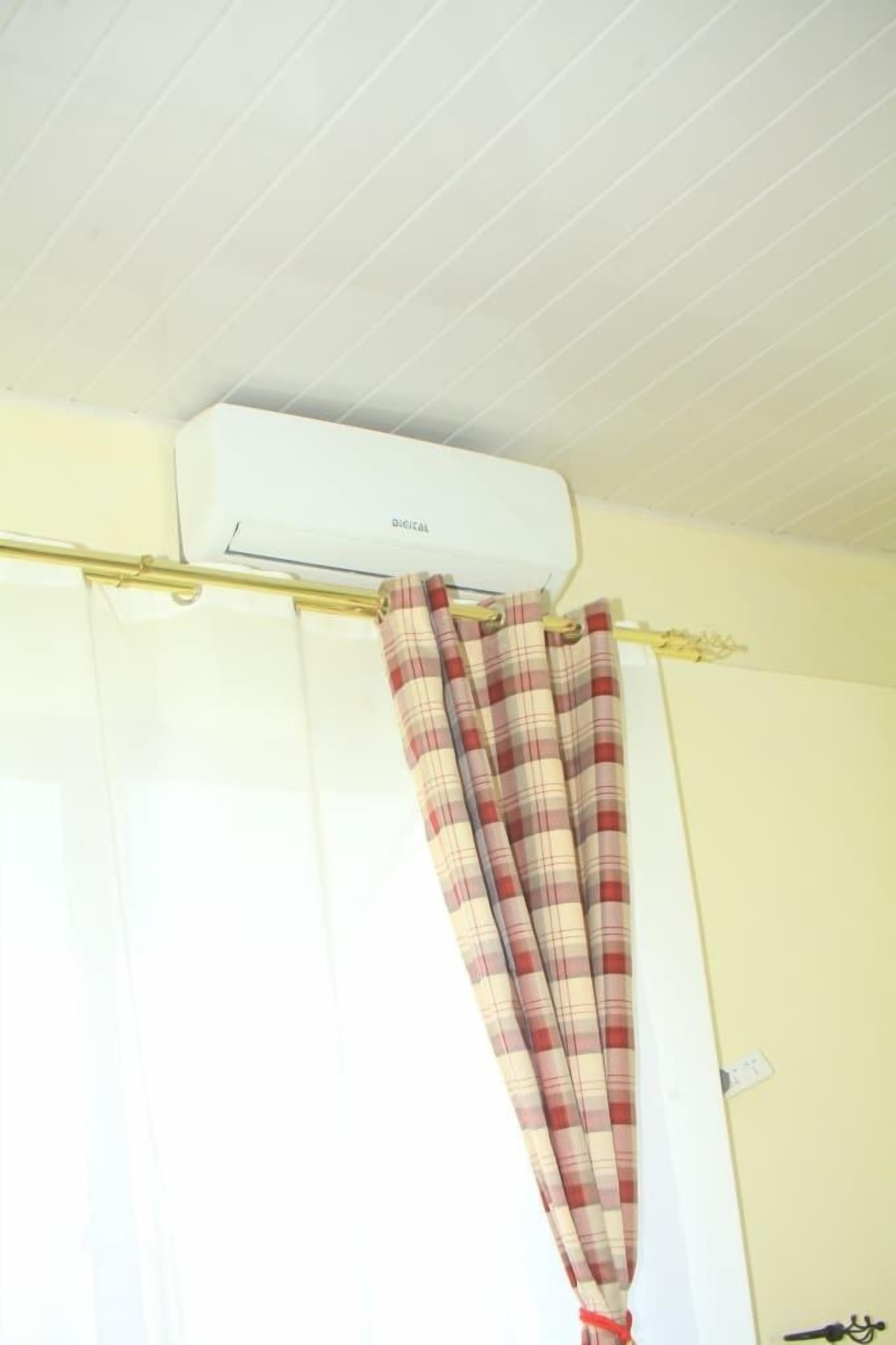 A room with a white air conditioning unit mounted on the ceiling, a plaid curtain hanging on a curtain rod, and a light-colored wall.