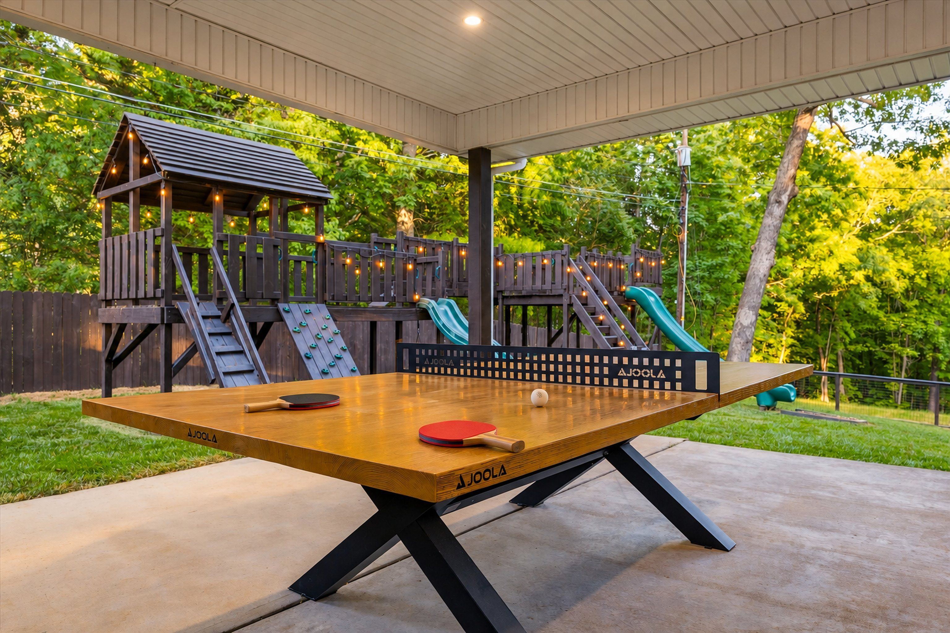 An outdoor ping pong table with a wooden surface and black legs, surrounded by a wooden deck and a play structure with slides.