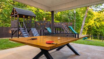 An outdoor ping pong table with a wooden surface and black legs, surrounded by a wooden deck and a play structure with slides.
