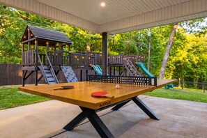 An outdoor ping pong table with a wooden surface and black legs, surrounded by a wooden deck and a play structure with slides. - River Views | A-Frame + Hot Tub + Fenced Yard + Adventure Fort + Fire Pit (Mount Airy)