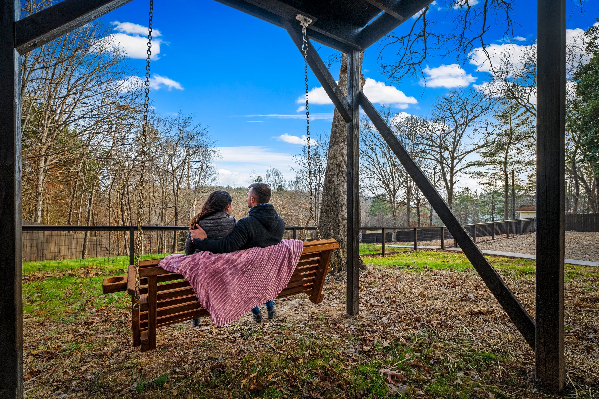 A wooden swing bench with a pink blanket, set in a park with trees and a clear sky.