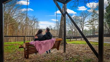 A wooden swing bench with a pink blanket, set in a park with trees and a clear sky.