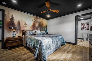 A bedroom with a bed, a nightstand, a ceiling fan, a wall mural, and a bathroom visible through an open door. - River Views | A-Frame + Hot Tub + Fenced Yard + Adventure Fort + Fire Pit (Mount Airy)