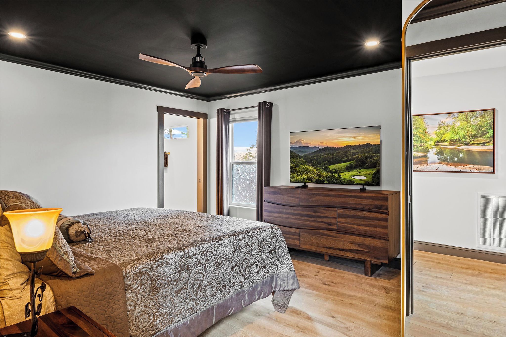 A bedroom with a bed, a nightstand, a dresser, a television, and a window with curtains.