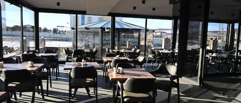 Un restaurante moderno con amplios ventanales, mesas al aire libre y vistas a una obra en construcción.