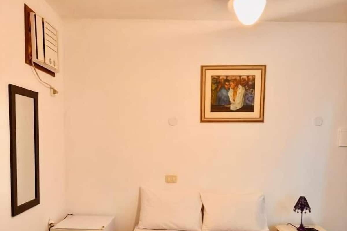 A hotel room with a bed, a nightstand, a ceiling fan, a wall-mounted painting, and a small cabinet.