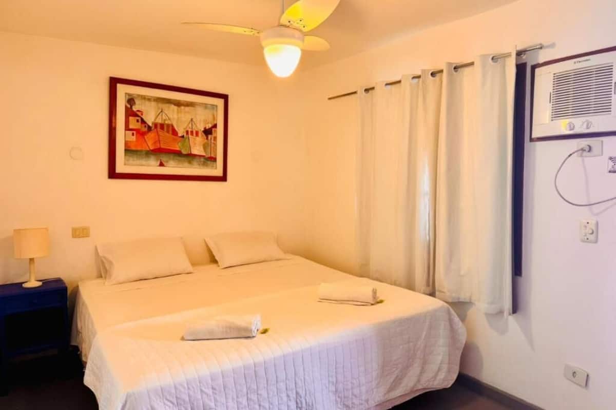 A hotel room with a bed, a ceiling fan, a painting on the wall, and a window with curtains.